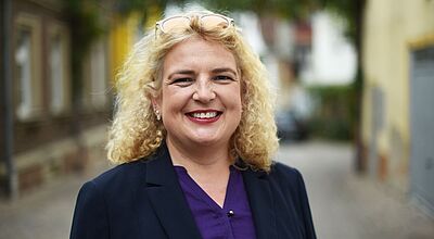 Anke Budde wurde in Berlin als ASR-Präsidentin wiedergewählt Eine Frau mit lockigem blondem Haar steht lächelnd in einer Gasse im Freien. Sie trägt ein dunkelblaues Jackett und eine violette Bluse.