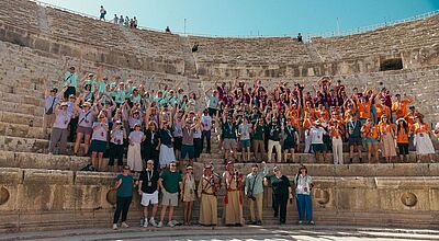 Der diesjährige GX-Gipfel fand in Jordanien statt, im nächsten Jahr geht es nach Marokko Eine große Gruppe von Menschen posiert auf den Stufen eines antiken Amphitheaters bei sonnigem Wetter. Die Teilnehmenden tragen bunte, farblich abgestimmte T-Shirts in Gruppenfarben (Lila, Türkis, Orange, Dunkelgrün) und heben jubelnd die Arme. Vorne stehen einige Personen in traditioneller Kleidung. Der Hintergrund zeigt die steinernen Sitzreihen des Theaters unter einem klaren blauen Himmel.