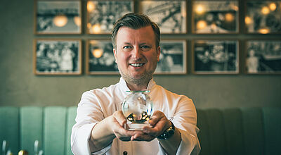 Sternekoch Kevin Fehling im The Globe an Bord der Europa. Sternekoch Kevin Fehling in weißer Uniform präsentiert eine Kristalltrophäe, im Hintergrund sind alte Fotografien und eine grüne Restauranteinrichtung zu erkennen.