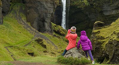 In Island gibt es für Kinder jede Menge zu entdecken. Foto: AscentXmedia/iStock Zwei Mädchen in Rückansicht mit Mützen und Winterjacken schauen auf einen Wasserfall, der von grün bewachsenen Felsen herunterstürzt.